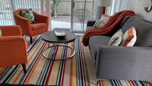 Ein Wohnzimmer mit grauem Sofa, einem orangefarbenen Sessel und einem kleinen runden Tisch mit Tablett. Ein farbenfroher Streifen Teppich bedeckt den Boden.