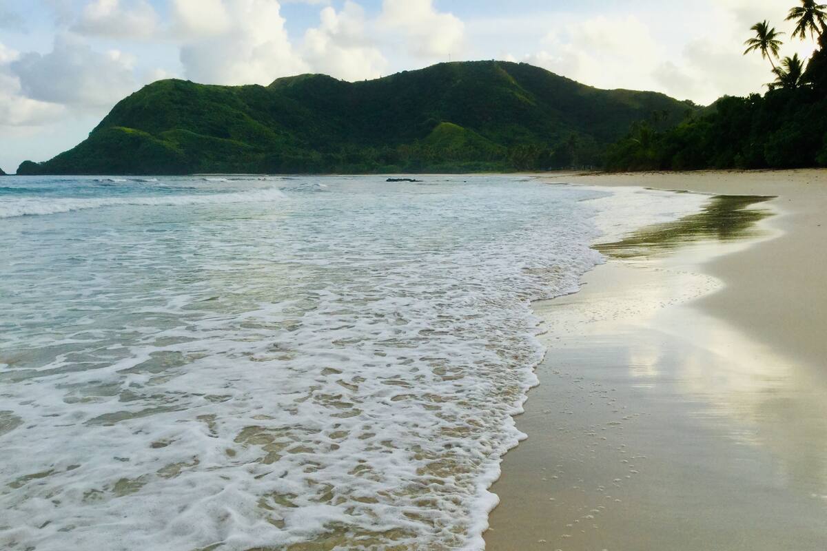 A beach with gentle waves, a sandy shore, and a mountain in the background.