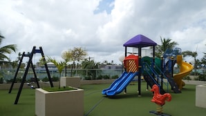 Un parque infantil con toboganes y columpios sobre césped artificial, rodeado de palmeras y un cielo nublado.
