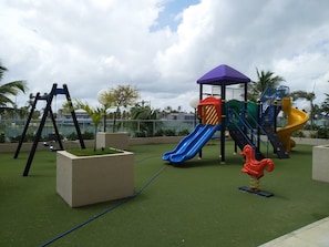 Ein Spielplatz mit Rutschen und Schaukeln auf Kunstrasen, umgeben von Palmen und einem bewölkten Himmel.