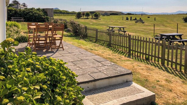 A patio with a wooden table and chairs, a stone pathway, a wooden fence, and a grassy field with picnic tables. - 4 bedroom accommodation in Criccieth, near Porthmadog (Criccieth, near Porthmadog)