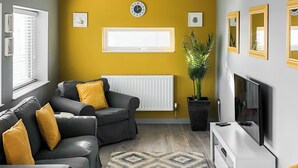 A living room with a sofa, a TV, a radiator, a plant, and framed pictures on the wall. - 4 bedroom accommodation in Criccieth, near Porthmadog (Criccieth, near Porthmadog)