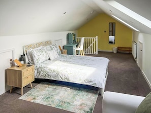 Une chambre avec un plafond en pente, un lit avec une literie à motifs floraux, un bureau avec une chaise, un radiateur et une fenêtre.