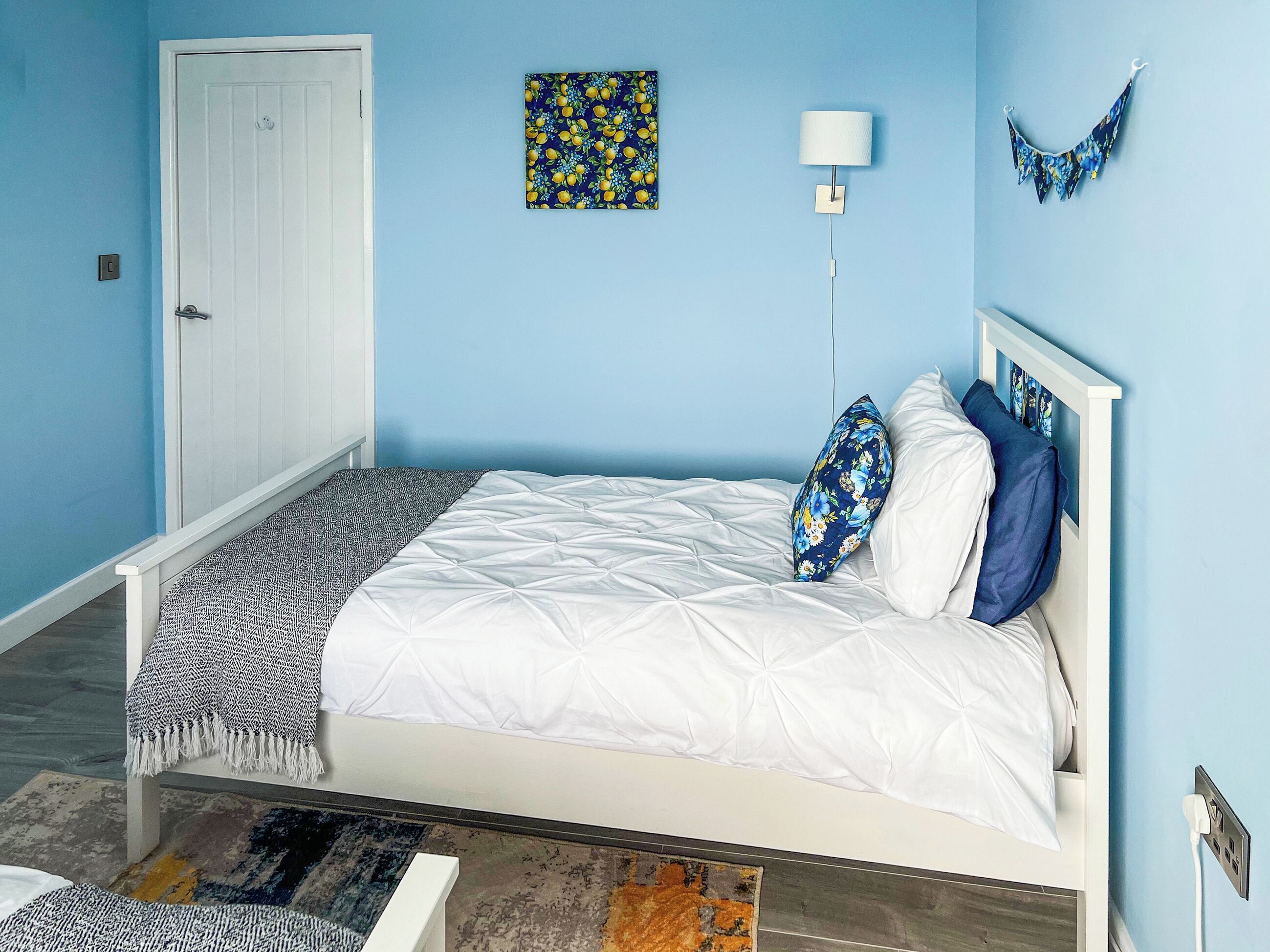 A bedroom with a white bed, a patterned throw blanket, a blue and yellow wall art, a white bedside lamp, and a blue and yellow curtain.