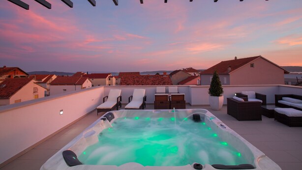 Een terras op het dak met een jacuzzi, lounge fauteuils en uitzicht op de stad tijdens de zonsondergang.