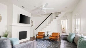 A modern living room with a grey sofa, two armchairs, a coffee table, and a patterned rug. A flat-screen TV is mounted on the wall, and a ceiling fan is visible. - Spacious, Historic Home with Porches (Charleston)