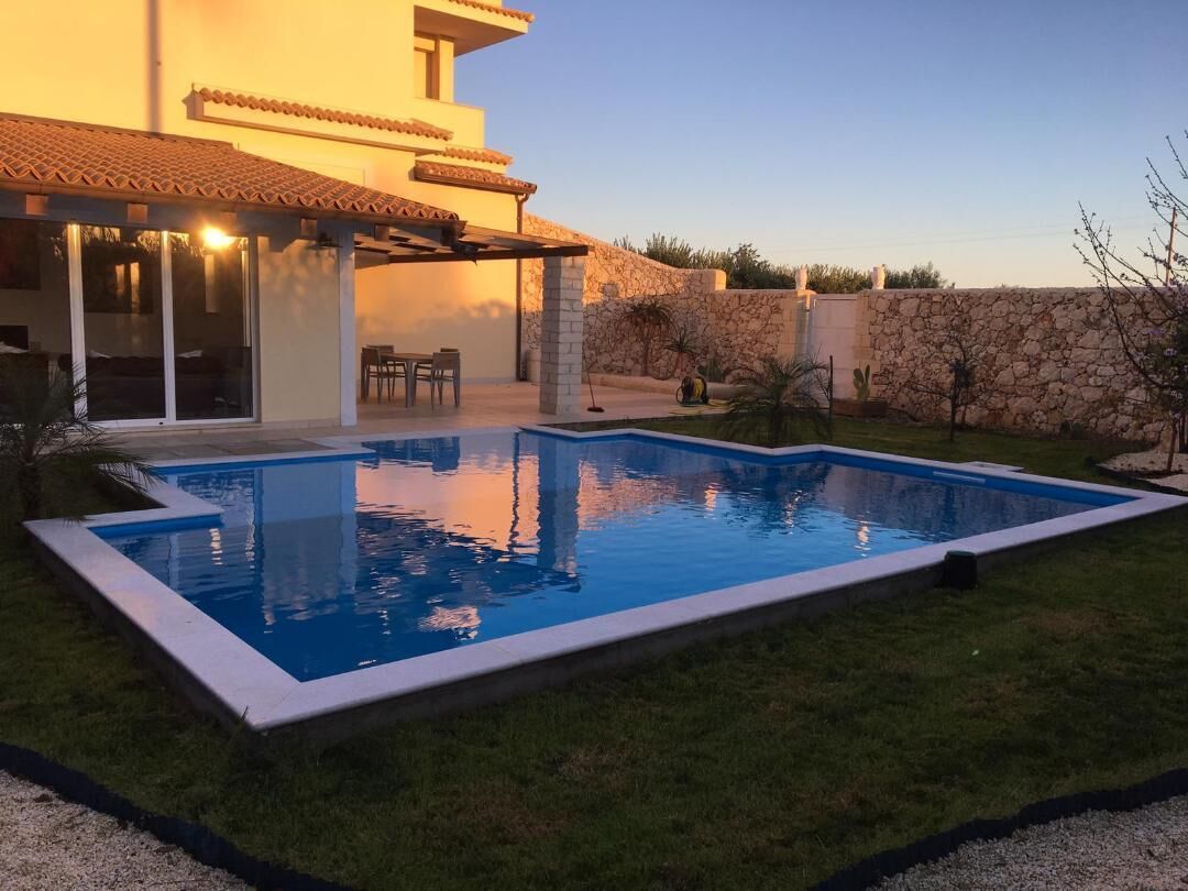 A rectangular swimming pool with a stone wall and a patio area with chairs and a table.