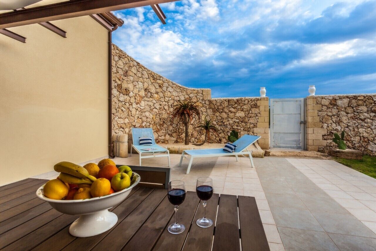 Une terrasse extérieure avec un mur en pierre, deux fauteuils de jardin, une table avec un panier de fruits et des verres à vin, et un ciel dégagé.