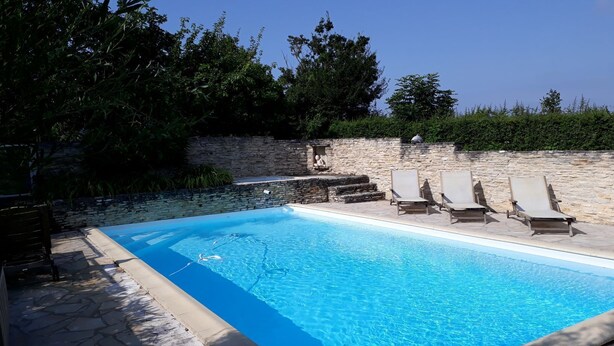 A swimming pool with stone tiles and a stone wall. There are lounge chairs and a stone bench.