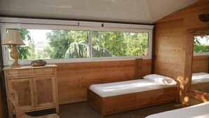 A wooden cabin room with two beds, a wooden cabinet, a woven chair, and a large window with a view of trees.