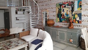 A living room with a white sofa, a wicker chair, a glass coffee table, a white cabinet, a spiral staircase, and a colorful abstract painting on the wall.
