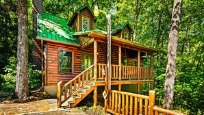 A wooden cabin with a green roof, a porch, and a staircase leading to the main entrance.