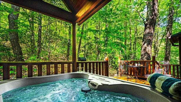 A hot tub in a forested area with a wooden deck and outdoor furniture.