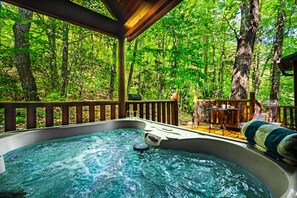 Un jacuzzi en un área boscosa con terraza de madera y mobiliario de exterior.