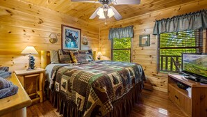 A cozy cabin room with a wooden interior, a bed with a patterned quilt, a ceiling fan, a television, and a window with a view of greenery.