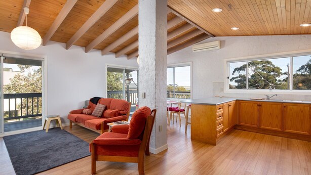 Uma sala de estar ampla com teto de madeira, um sofá, uma poltrona e uma cozinha com armários de madeira e uma janela grande.