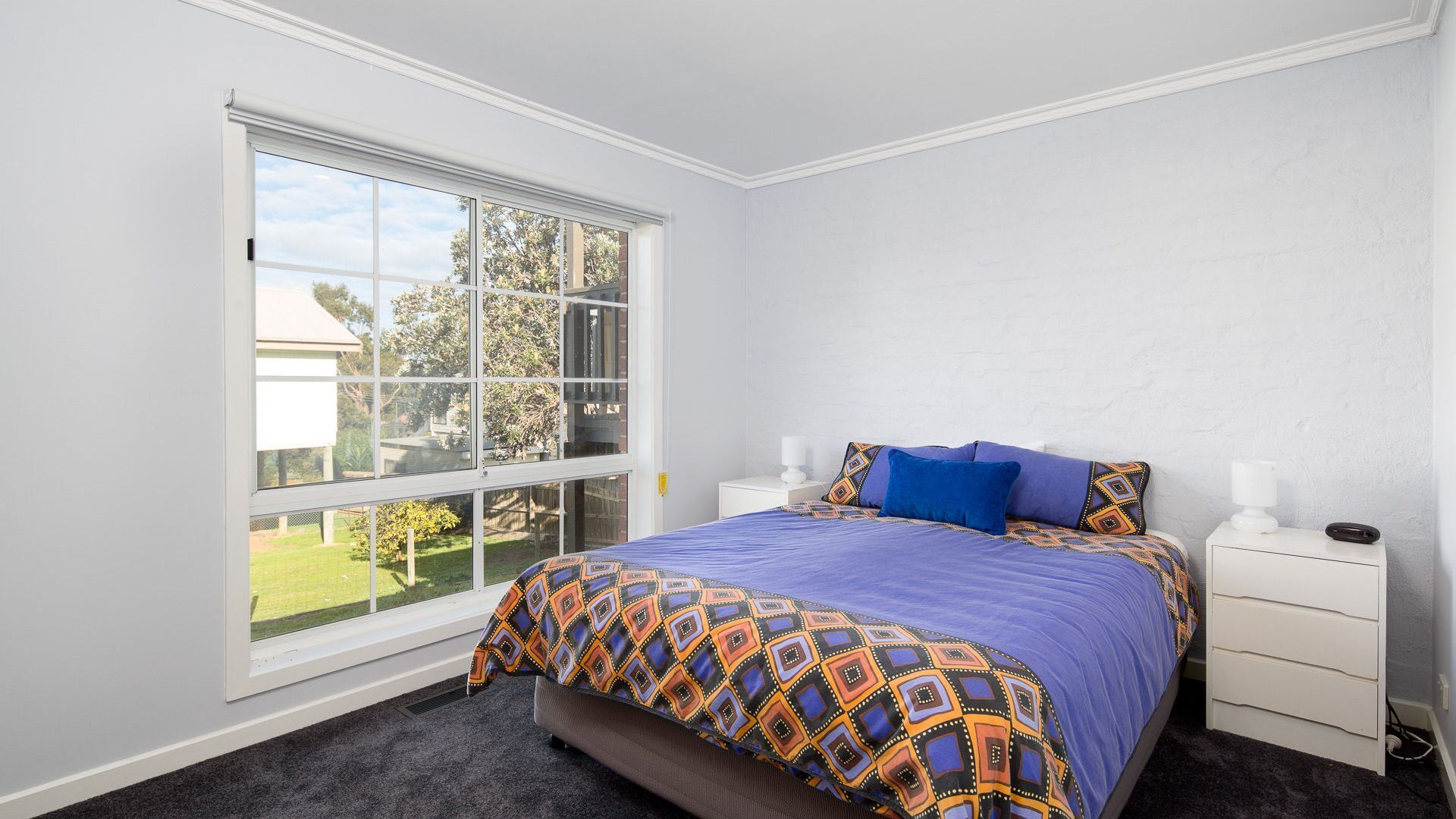 A bedroom with a large window, a bed with patterned bedding, a bedside table, and a nightstand.