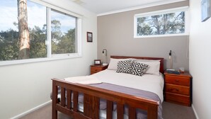 Une chambre avec un lit en bois, deux tables de chevet, une commode, une fenêtre donnant sur des arbres et un tableau encadré accroché au mur.