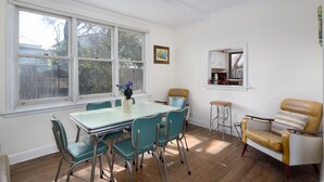 Un coin repas avec une table et des chaises, une fenêtre donnant sur des arbres, un petit tabouret et un fauteuil ancien.