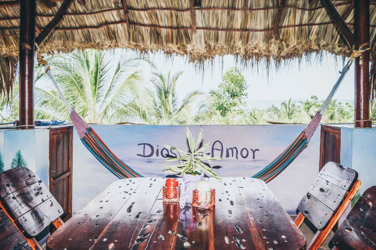 A wooden table with chairs, a thatched roof, and a mural with the words "Dios Amor".