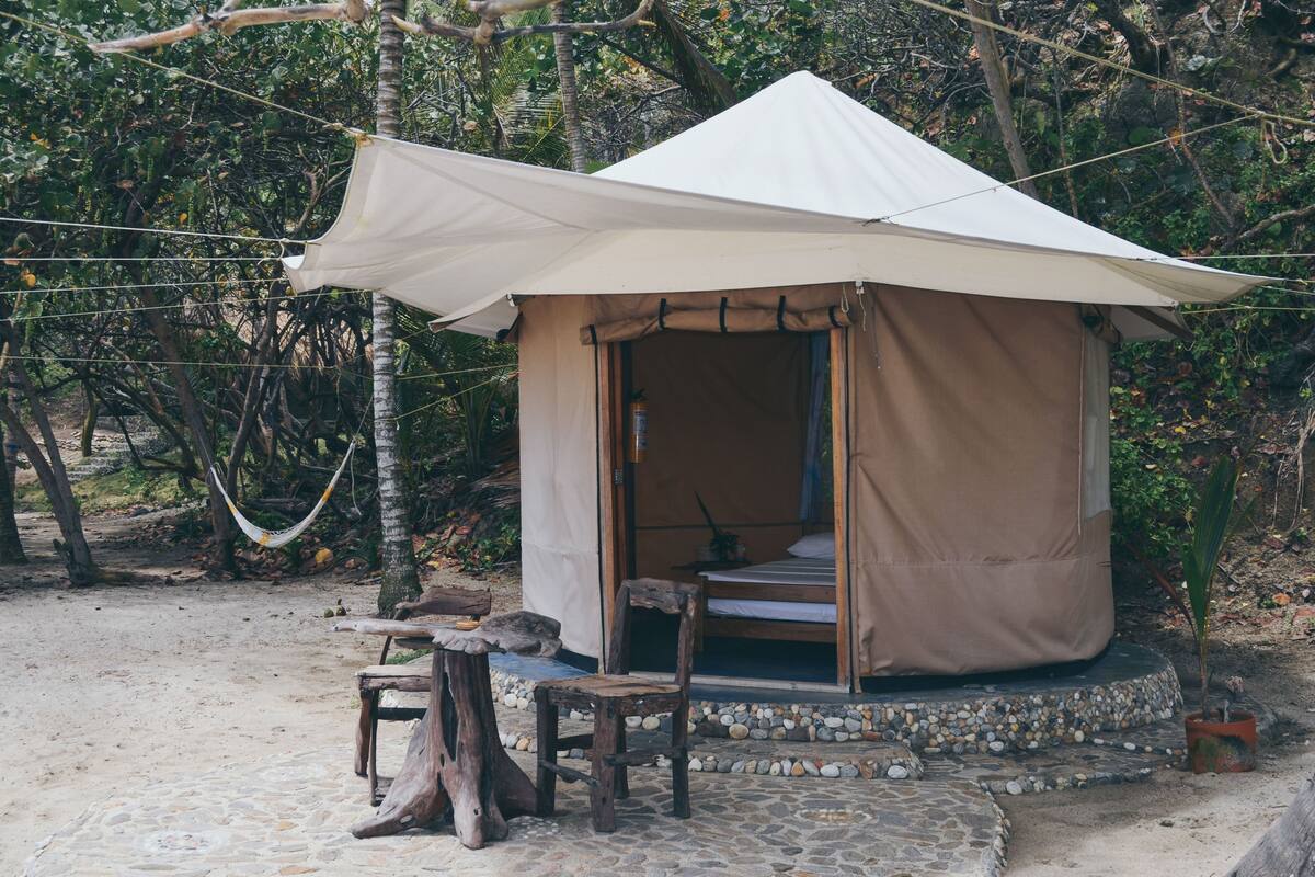 A small, single-room tent with a bed, a table, and a chair, surrounded by natural vegetation and a hammock.