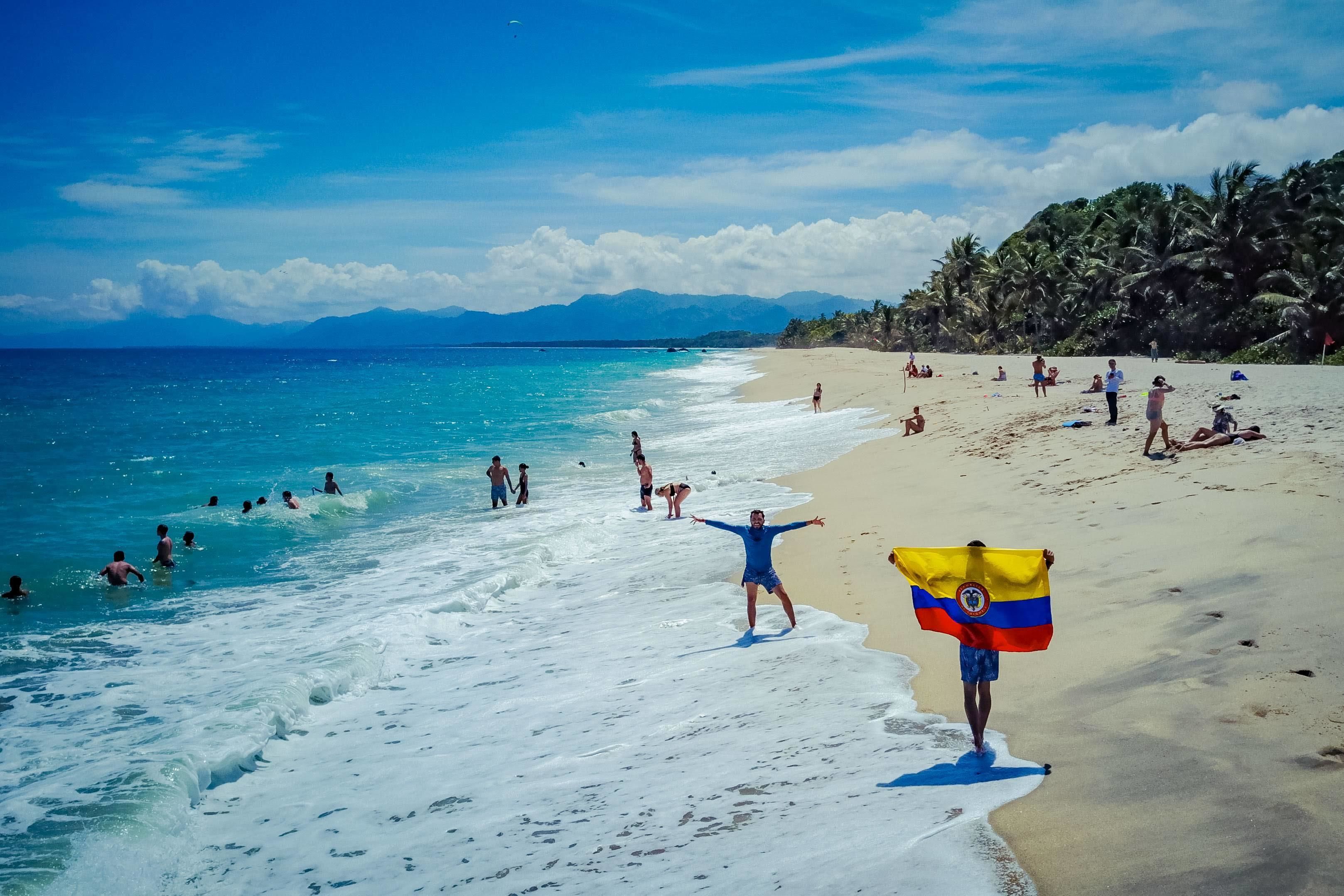 Escena de playa con gente nadando y caminando por la orilla, palmeras al fondo y montañas a lo lejos.