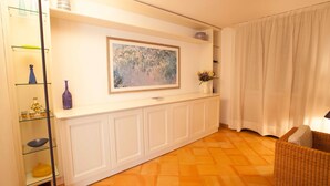 Una habitación con una consola blanca, un cuadro enmarco, un sillón de mimbre y una estantería de cristal con adornos.