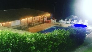 A nighttime view of a hotel with a swimming pool, lounge chairs, and a building with string lights. - Chácara Vovó Joana (Atibaia)