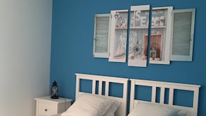Un dormitorio con pared azul, cama blanca con dos almohadas, mesita de noche con lámpara y estante de pared con objetos decorativos.