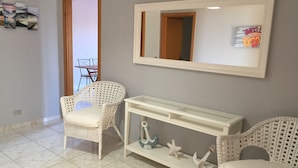 Una habitación con dos sillones de mimbre, una consola blanca con adornos, un espejo grande y una puerta que da a otra estancia.