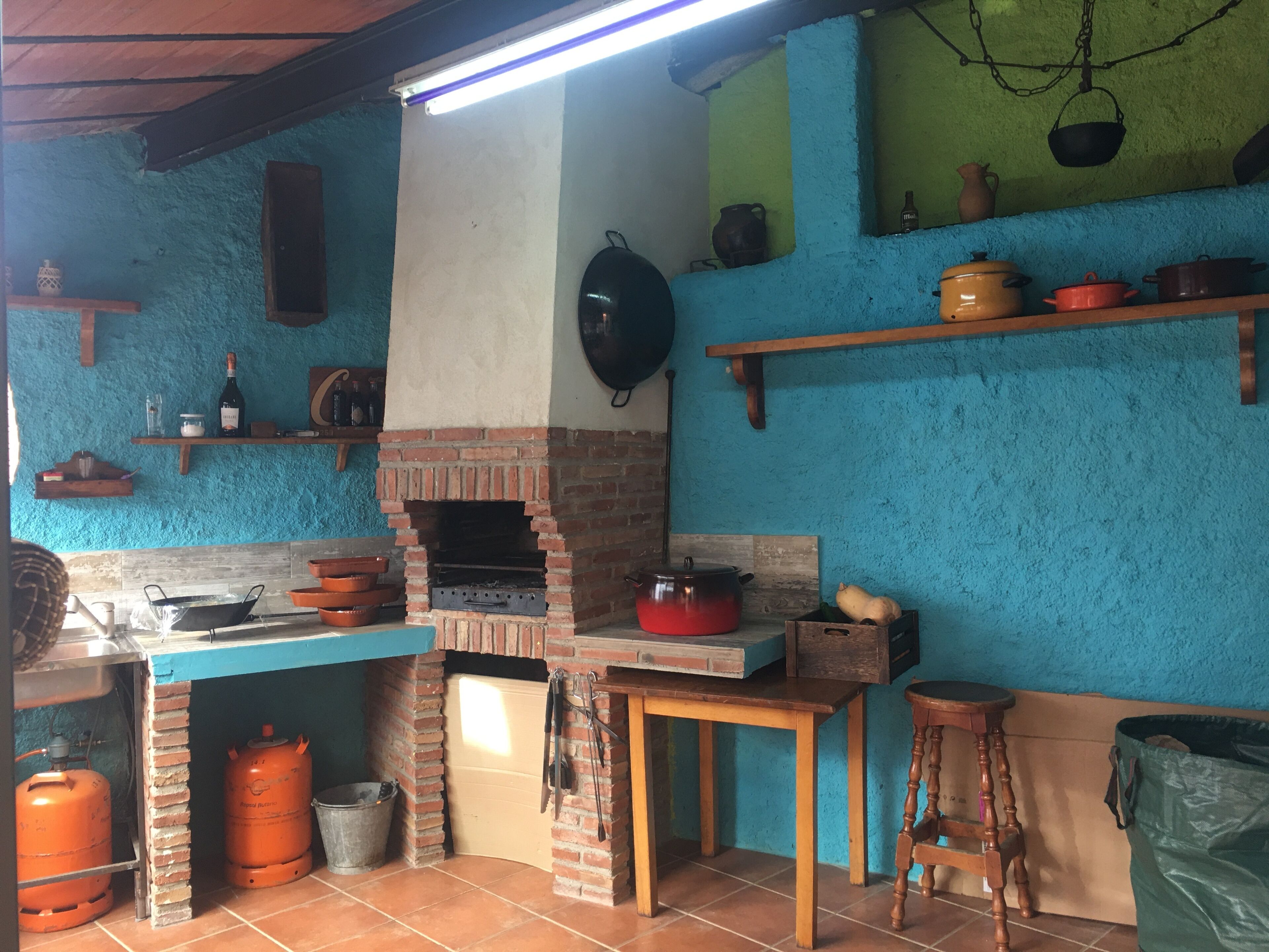 A kitchen with a brick oven, shelves with pots, a wooden table, and gas cylinders.