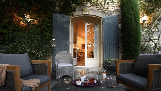 A cozy outdoor seating area with wicker furniture, a table set with snacks and drinks, and a stone building entrance in the background. - Welcome to La Maison du Bonheur (Gordes)