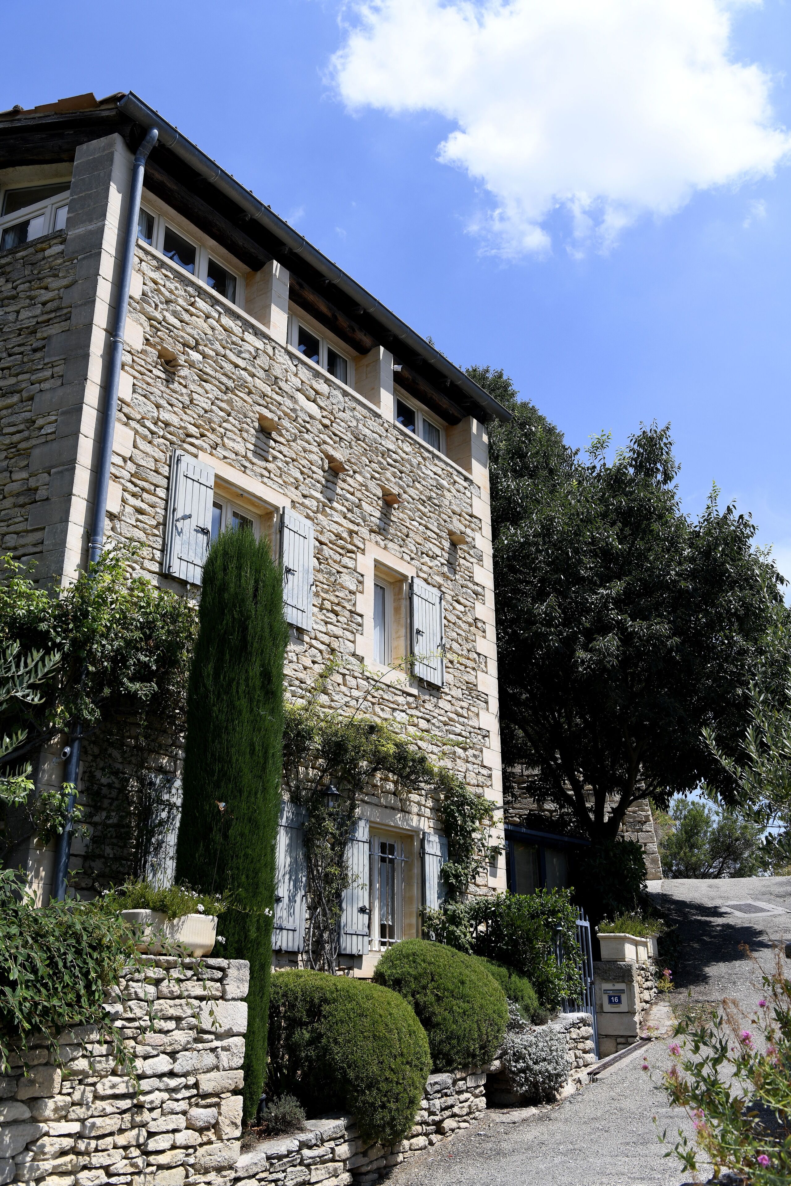 Un edificio de piedra con fachada tradicional, múltiples ventanas con contraventanas y un jardín bien cuidado con diversos arbustos y un conífero alto.