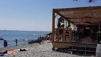 A beach with a wooden structure, possibly a restaurant or bar, and a rocky shoreline with people and umbrellas.