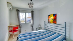 Un dormitorio con cama, escritorio, silla, ventana con cortinas, un cuadro en la pared y una araña.