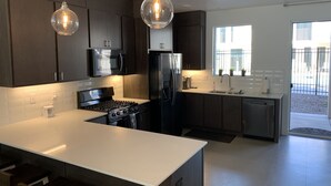 Eine moderne Küche mit dunklen Schränken, einer weißen Arbeitsplatte, Edelstahlgeräten, Pendelleuchten und Blick ins Freie durch große Fenster.