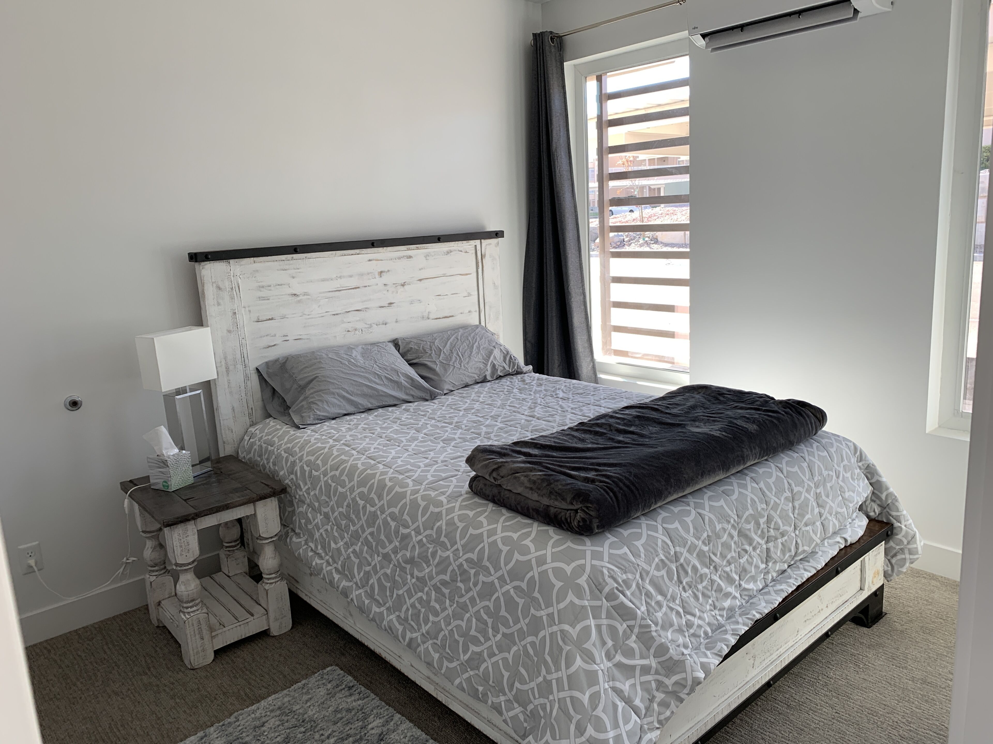 A bedroom with a white bed, grey patterned bedding, a bedside table with a lamp, and a view of the outdoors through a window.
