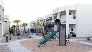 Ein Spielplatz mit Rutsche und Klettergerüst, umgeben von Sand und Palmen, mit modernen Gebäuden im Hintergrund.