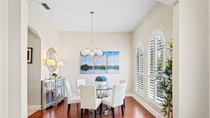 A dining area with a round table, white chairs, a chandelier, and a view through arched windows. - Homestead Beech: Private Pool, Golf, Water Park, Near Disney (Kissimmee)