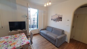 A living room with a grey sofa, a TV on a stand, a table with a floral tablecloth, and a window with a view of buildings. - BEST VIEW AND SUNNY Cà DEL SOL IT027042C2W4RXWZRV (Venezia)