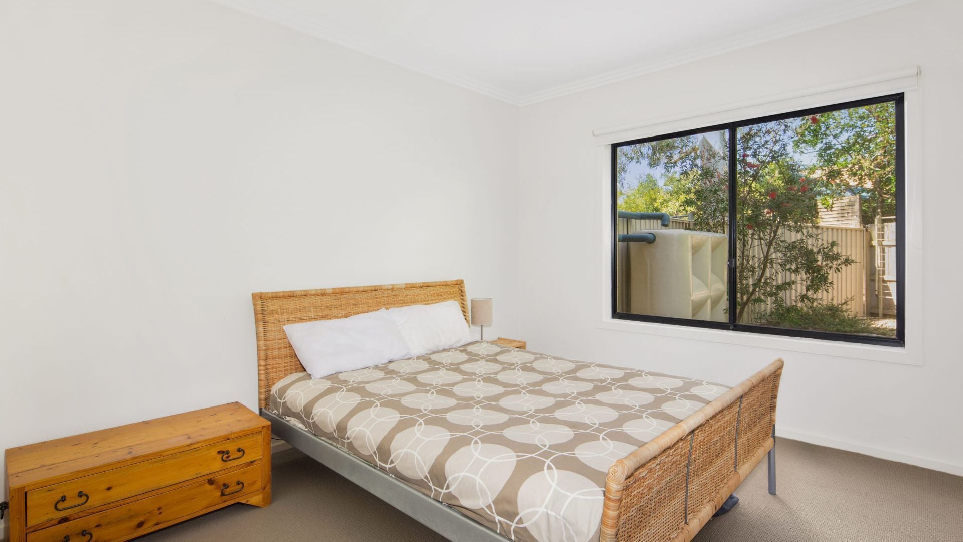 A bedroom with a bed, a wooden nightstand, a window with a view of the outside, and a white wall.