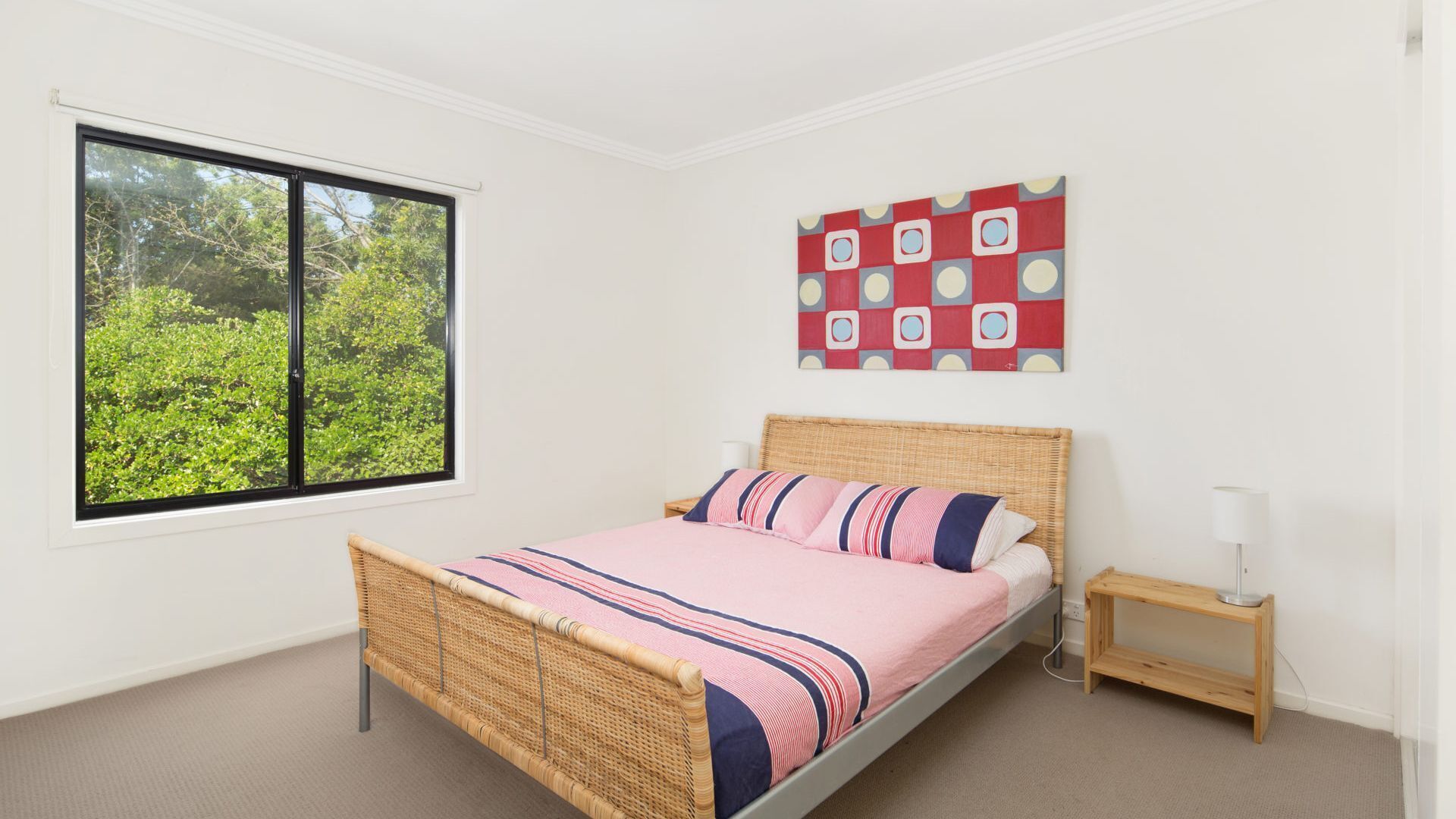 A bedroom with a bed, a window with a view of greenery, a bedside table, and a wall art piece.