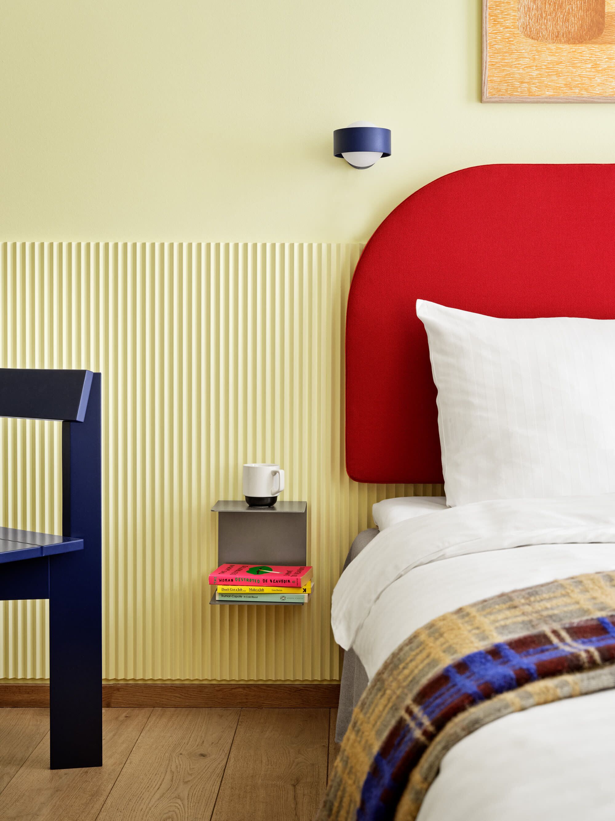 A neatly arranged hotel room with a bed, a bedside table with a cup and books, a chair, and a wall-mounted light fixture.