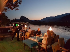 A lakeside dining area with tables and chairs, a waiter attending to guests, and a boat in the background.