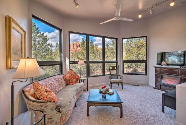 A living room with a sofa, coffee table, and TV, large windows with a view of trees and rocks, and a fireplace.