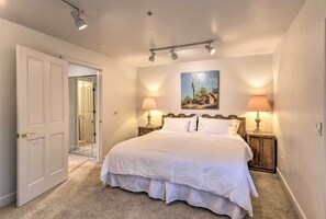 A bedroom with a large bed, two bedside lamps, a painting on the wall, and a door leading to another room.