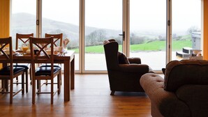 Un coin repas avec une table dressée pour quatre personnes, un canapé et un fauteuil placés face à une grande porte-fenêtre coulissante donnant sur un paysage verdoyant.