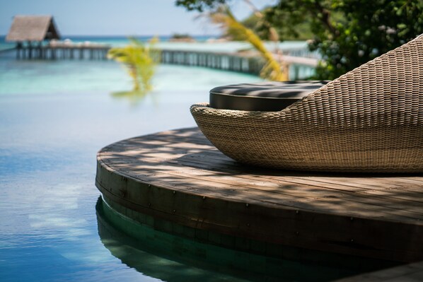 A wicker lounge chair by a pool with a wooden deck and a view of the ocean and overwater bungalows.