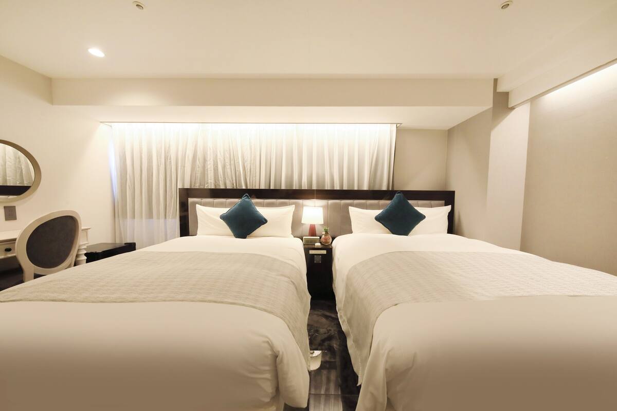 A hotel room with two beds, a desk, a chair, a nightstand with a lamp, and a large window with curtains.