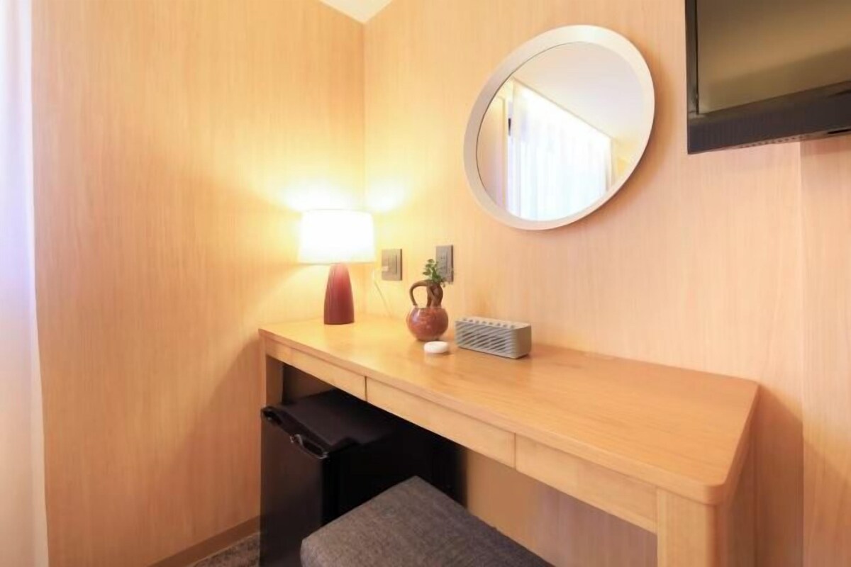 Ein Hotelzimmer mit einem Holztisch, einem runden Spiegel, einer Lampe, einer Vase und einem an der Wand befestigten Fernseher.